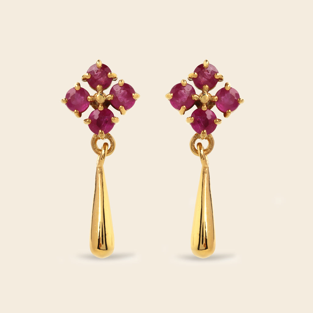 A pair of gold dangle earrings featuring a floral cluster of round-cut deep pinkish-red gemstones at the top and a polished gold teardrop-shaped drop below, set against a solid light beige background.