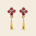 A pair of gold dangle earrings featuring a floral cluster of round-cut deep pinkish-red gemstones at the top and a polished gold teardrop-shaped drop below, set against a solid light beige background.