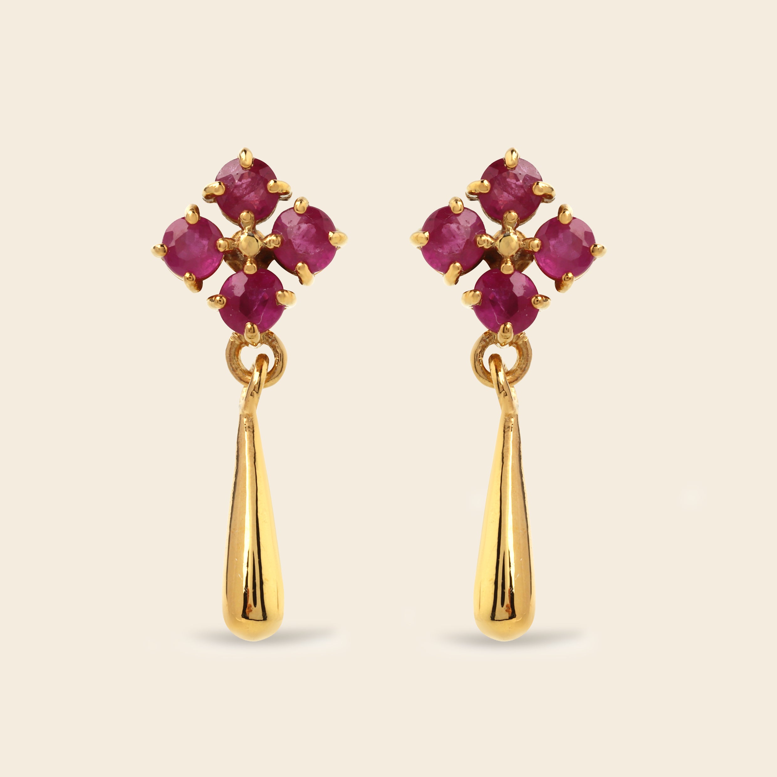 A pair of gold dangle earrings featuring a floral cluster of round-cut deep pinkish-red gemstones at the top and a polished gold teardrop-shaped drop below, set against a solid light beige background.