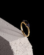A gold ring with a marquise-cut dark blue gemstone and smaller round blue stones, displayed on a textured white stone block against a solid black background.