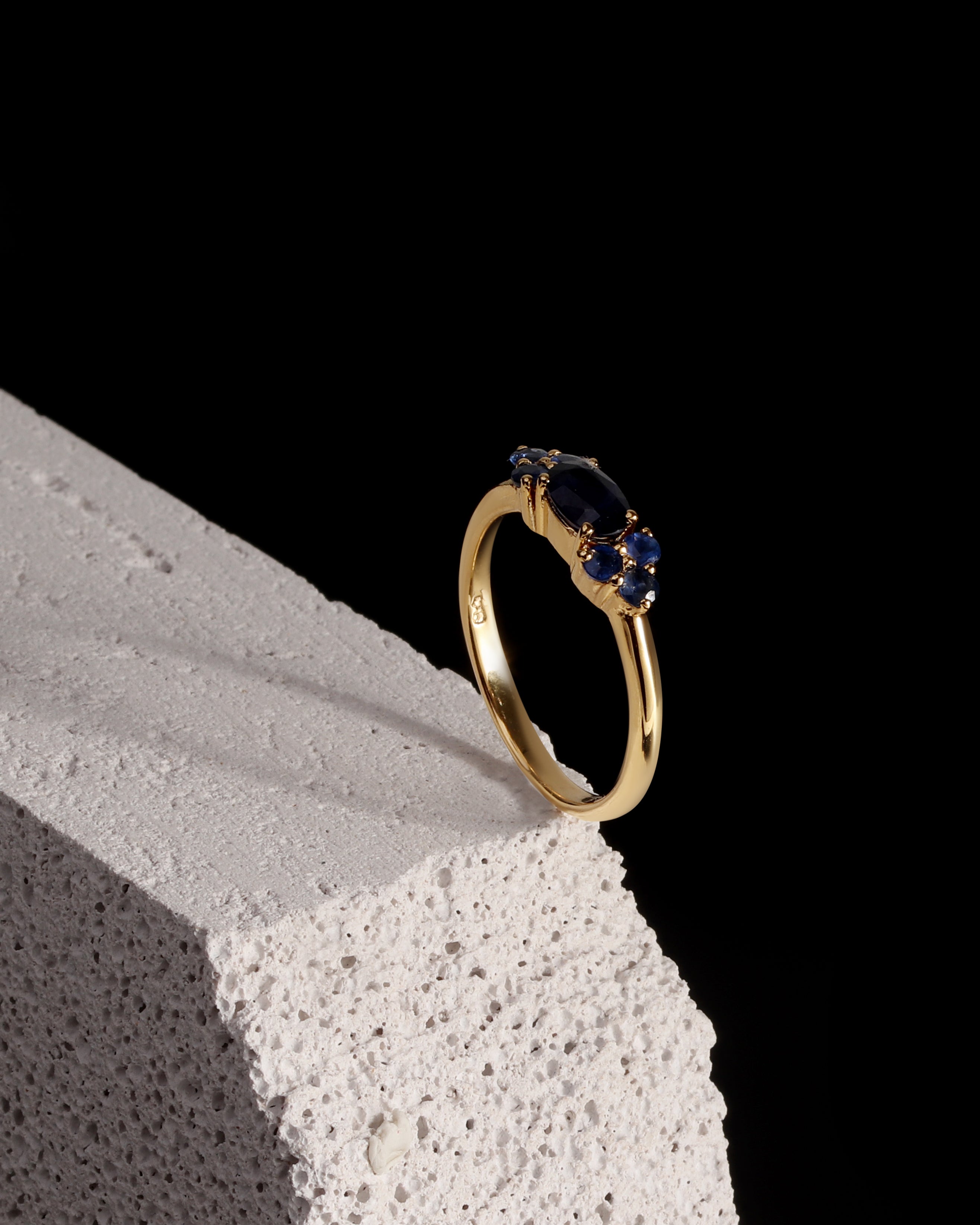 A gold ring with a marquise-cut dark blue gemstone and smaller round blue stones, displayed on a textured white stone block against a solid black background.