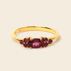 A yellow gold ring featuring a central oval-cut deep pinkish-red gemstone flanked by smaller round-cut dark red gemstones, set against a solid light beige background.