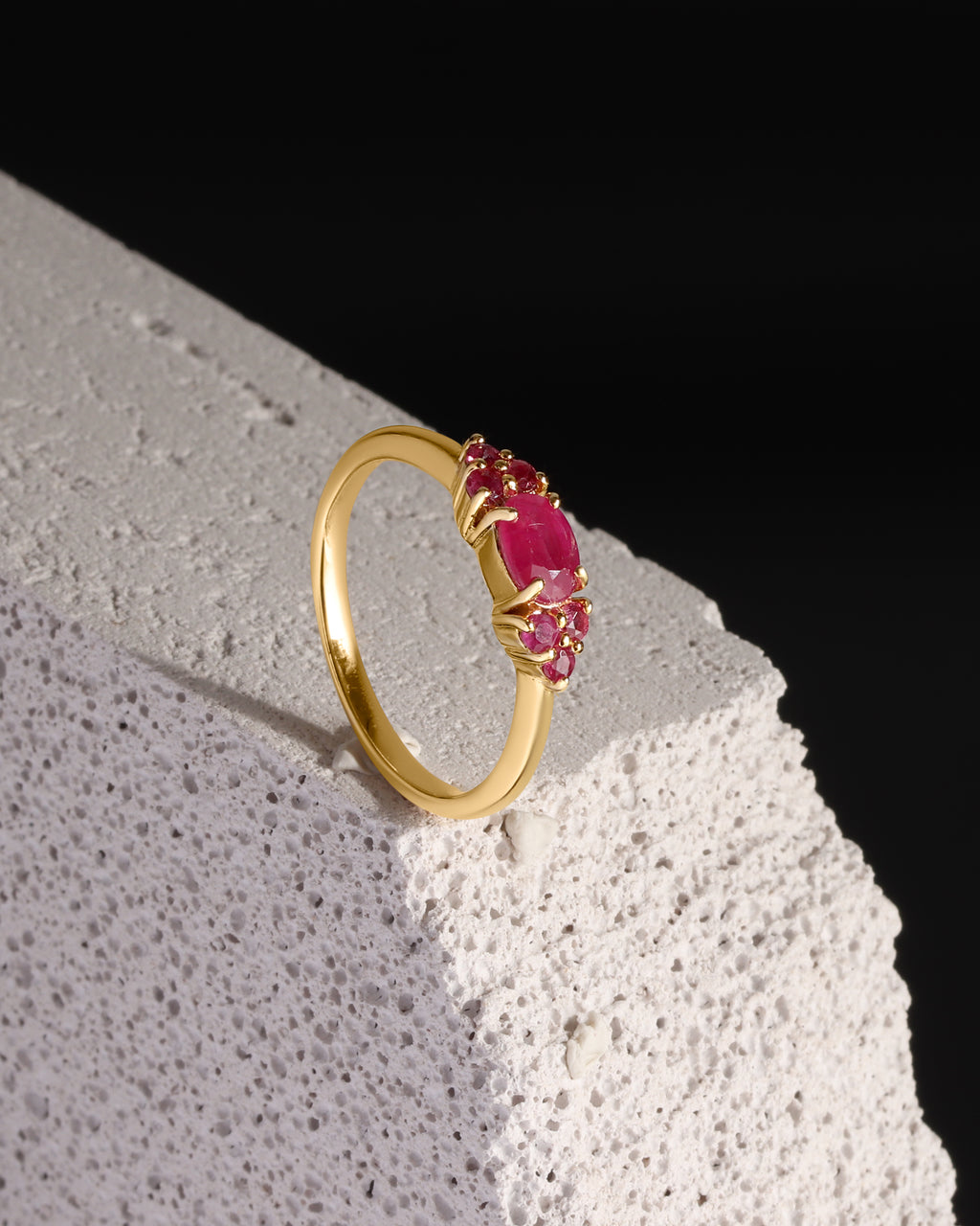 A gold ring with a cluster of oval pink gemstones, displayed on a textured white stone block against a solid black background.