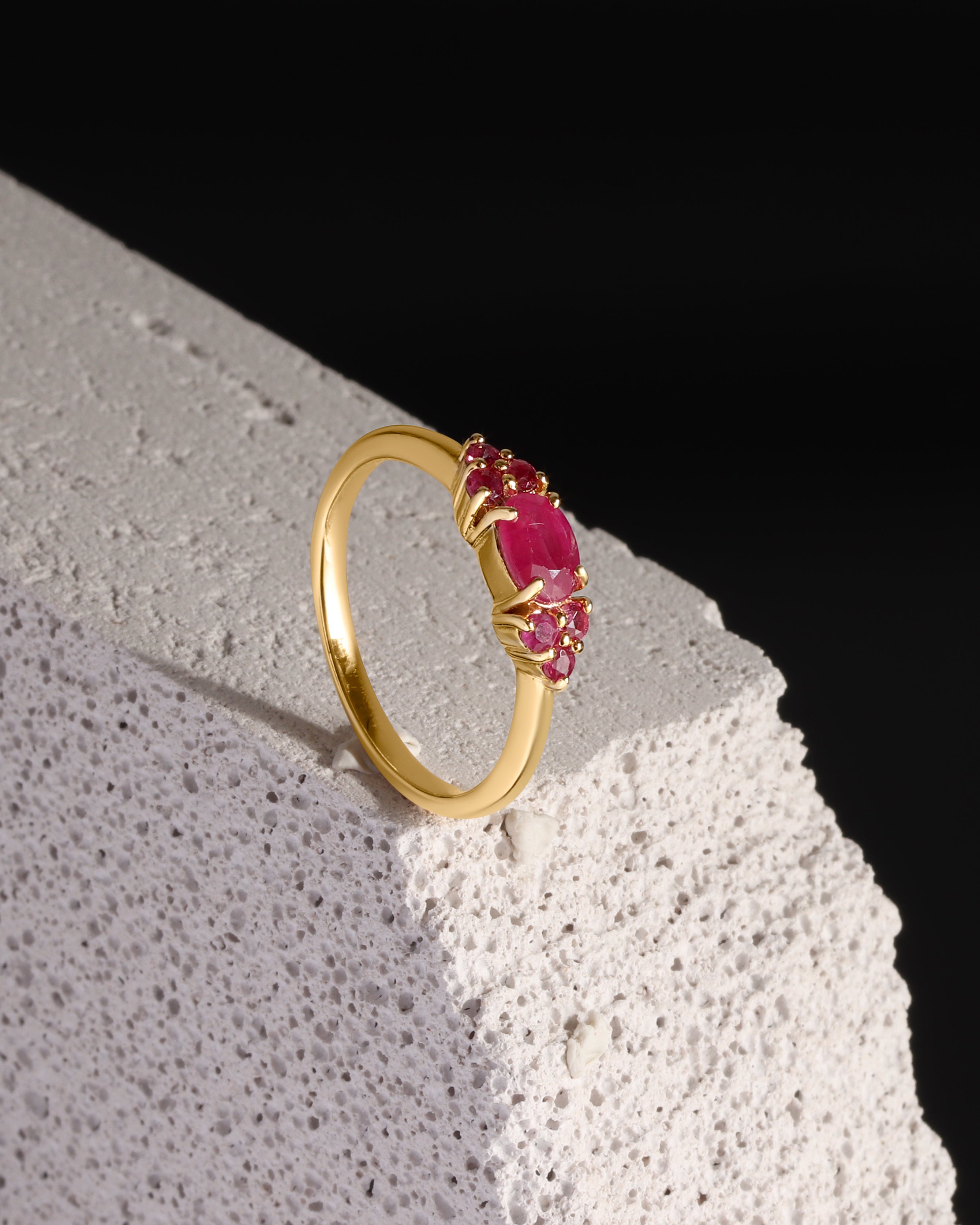 A gold ring with a cluster of oval pink gemstones, displayed on a textured white stone block against a solid black background.
