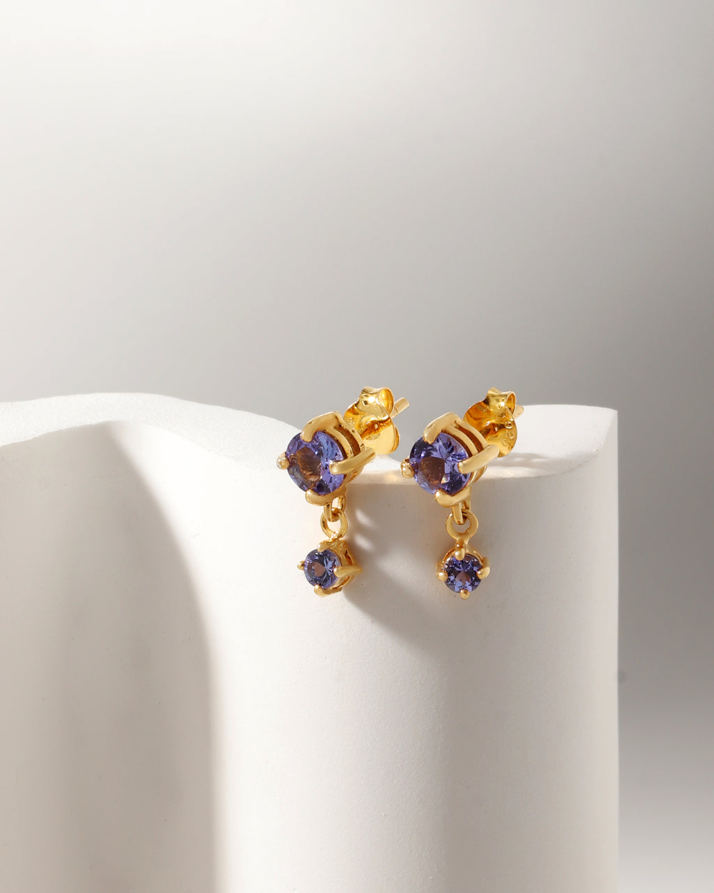 A pair of gold drop earrings with oval purple gemstones, displayed on a white curved stand against a light gray background.