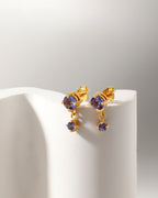 A pair of gold drop earrings with oval purple gemstones, displayed on a white curved stand against a light gray background.