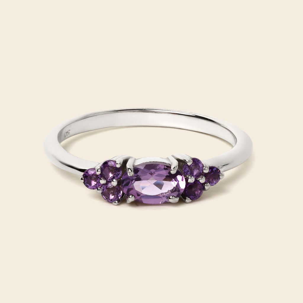 A silver ring featuring a central oval-cut purple gemstone flanked by smaller round purple gemstones, set against a solid light beige background.