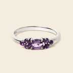 A silver ring featuring a central oval-cut purple gemstone flanked by smaller round purple gemstones, set against a solid light beige background.