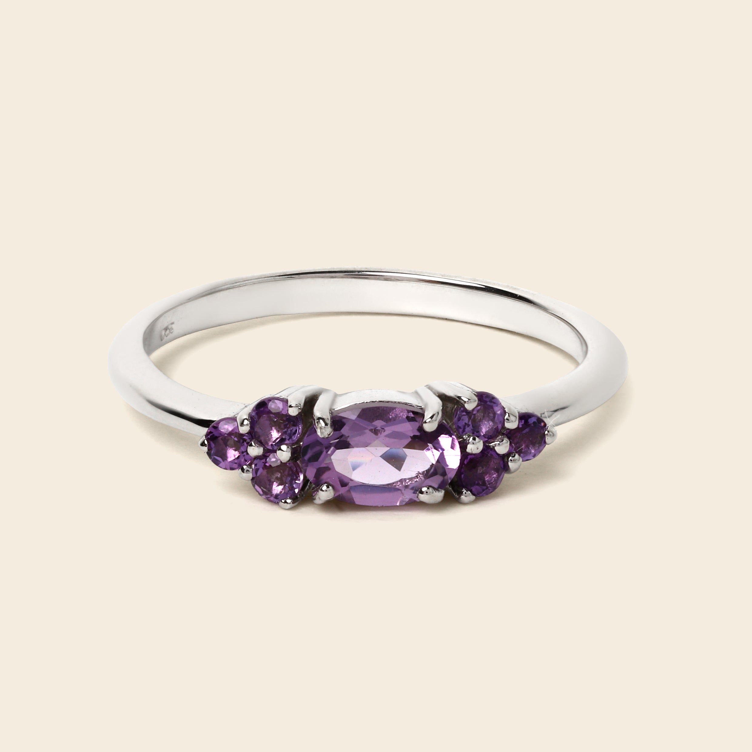 A silver ring featuring a central oval-cut purple gemstone flanked by smaller round purple gemstones, set against a solid light beige background.