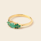 A gold ring featuring a oval-cut green emerald center stone and smaller round green stones on either side, set in a polished yellow gold band against a light beige background.