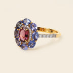 A yellow gold ring featuring a floral cluster setting with oval pinkish-red center stones, surrounded by blue and purple oval gemstones, and a pavé band with small clear stones, set against a solid cream background.