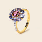 A yellow gold ring featuring a central oval pink gemstone surrounded by a halo of round blue gemstones and small clear accent stones on the band, set against a solid light beige background.