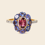 A yellow gold ring featuring a flower-shaped cluster with a central oval pinkish-red gemstone surrounded by round blue and oval brown gemstones, accented with small clear stones on the band, set against a solid beige background.