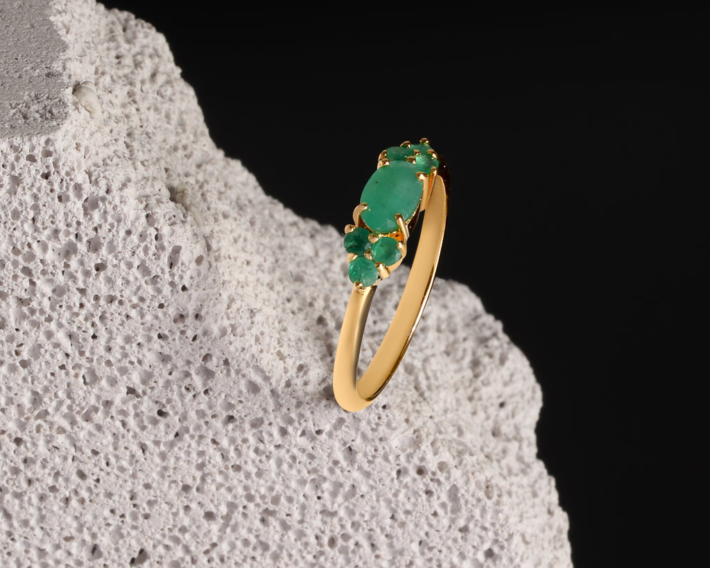 A gold ring with a cluster of green gemstones, displayed on a textured white stone against a solid black background.