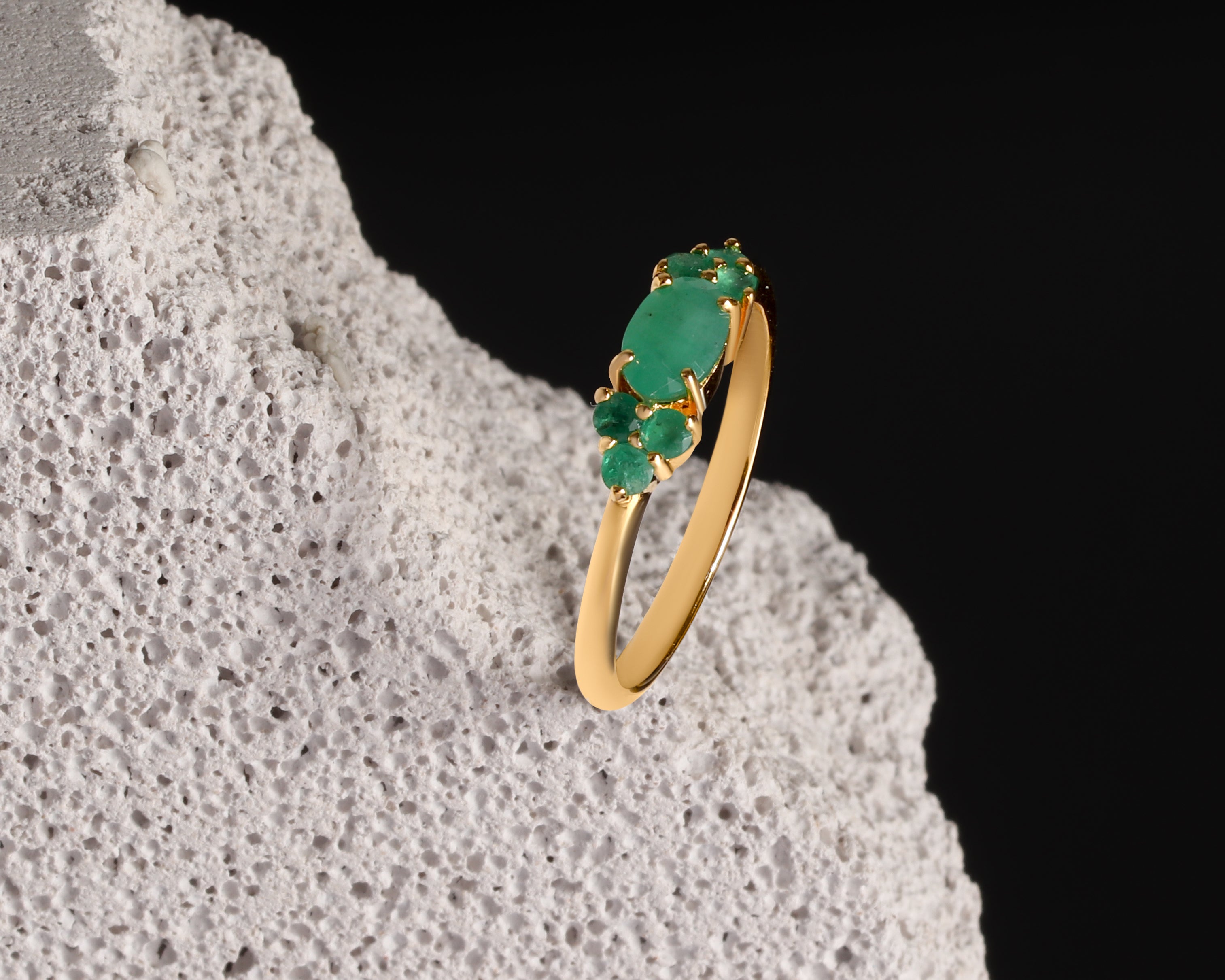 A gold ring with a cluster of green gemstones, displayed on a textured white stone against a solid black background.