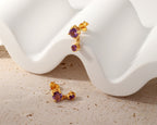 A pair of gold-toned stud earrings with oval purple gemstones, displayed on a white sculptural tray over a beige textured surface.
