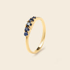 A gold band ring featuring a row of small round blue gemstones, photographed on a clean, solid, light beige background.