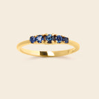 A yellow gold ring featuring five round-cut blue gemstones set in a row on a clean, light beige background.