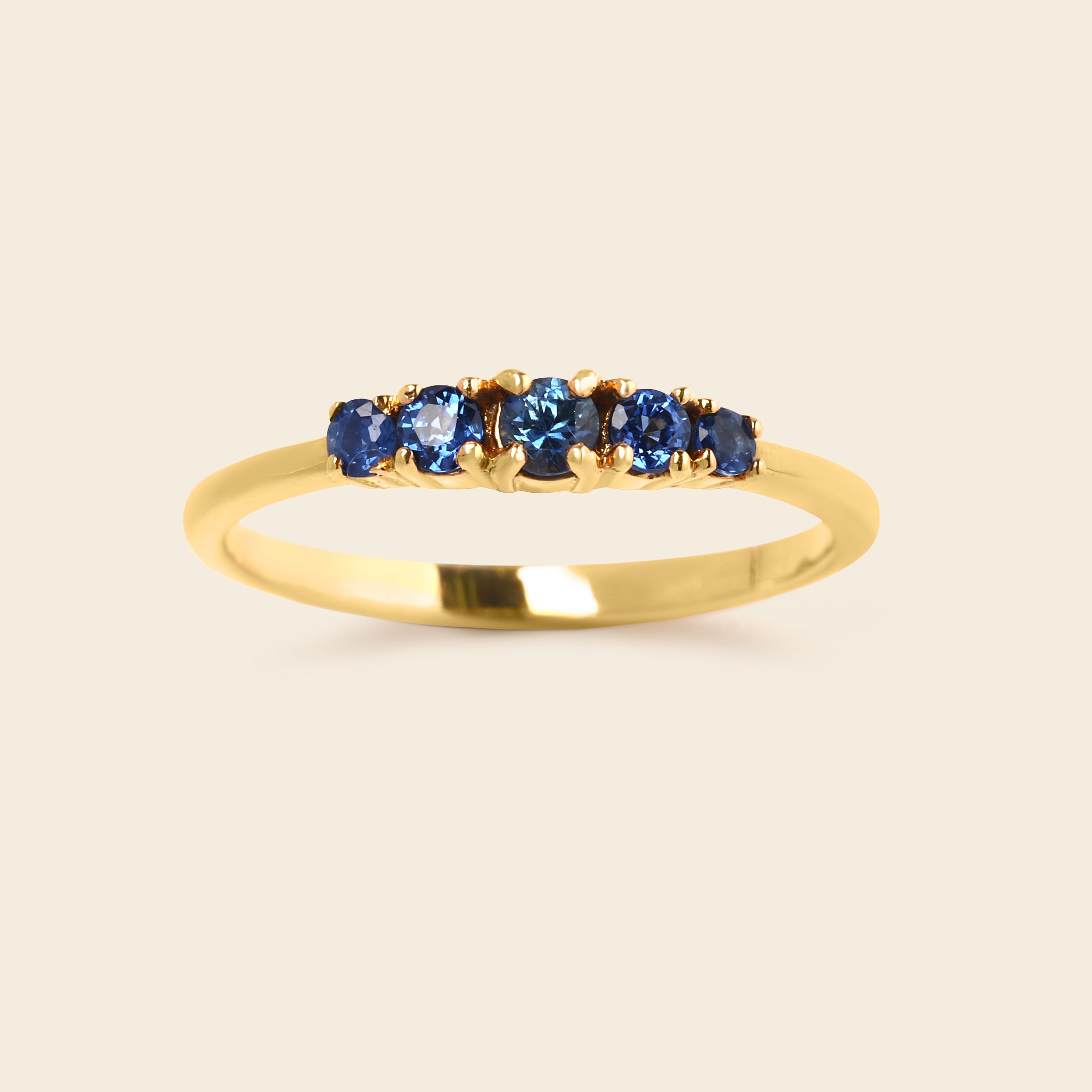 A yellow gold ring featuring five round-cut blue gemstones set in a row on a clean, light beige background.