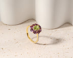 A gold ring with a flower-shaped cluster setting featuring a green peridot center stone and surrounding purple amethyst petals, accented with small clear stones on the band, set against a neutral beige textured surface with white ceramic props.