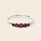A silver band ring featuring a row of five oval-cut deep red gemstones in a prong setting, presented on a solid light beige background.