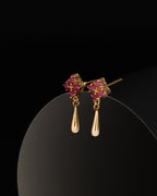 A pair of gold dangle earrings with pink gemstone clusters at the top and teardrop-shaped gold drops below, displayed on a black curved surface against a black background.