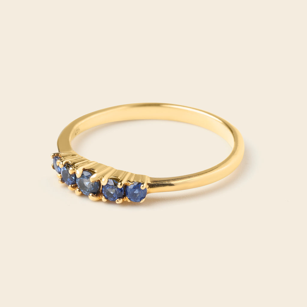 A gold ring featuring five small round-cut blue gemstones set in a curved row along the front, photographed on a solid light beige background.