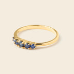 A gold ring featuring five small round-cut blue gemstones set in a curved row along the front, photographed on a solid light beige background.