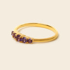 A gold ring featuring five small round purple gemstones set in a curved row, photographed on a plain light beige background.