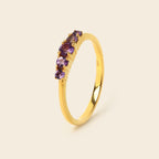 A yellow gold ring featuring a row of five purple amethyst gemstones, set against a solid light beige background.