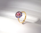 A yellow gold ring featuring a central oval pink gemstone surrounded by a halo of round blue gemstones and small clear accent stones on the band, displayed on a white curved surface.