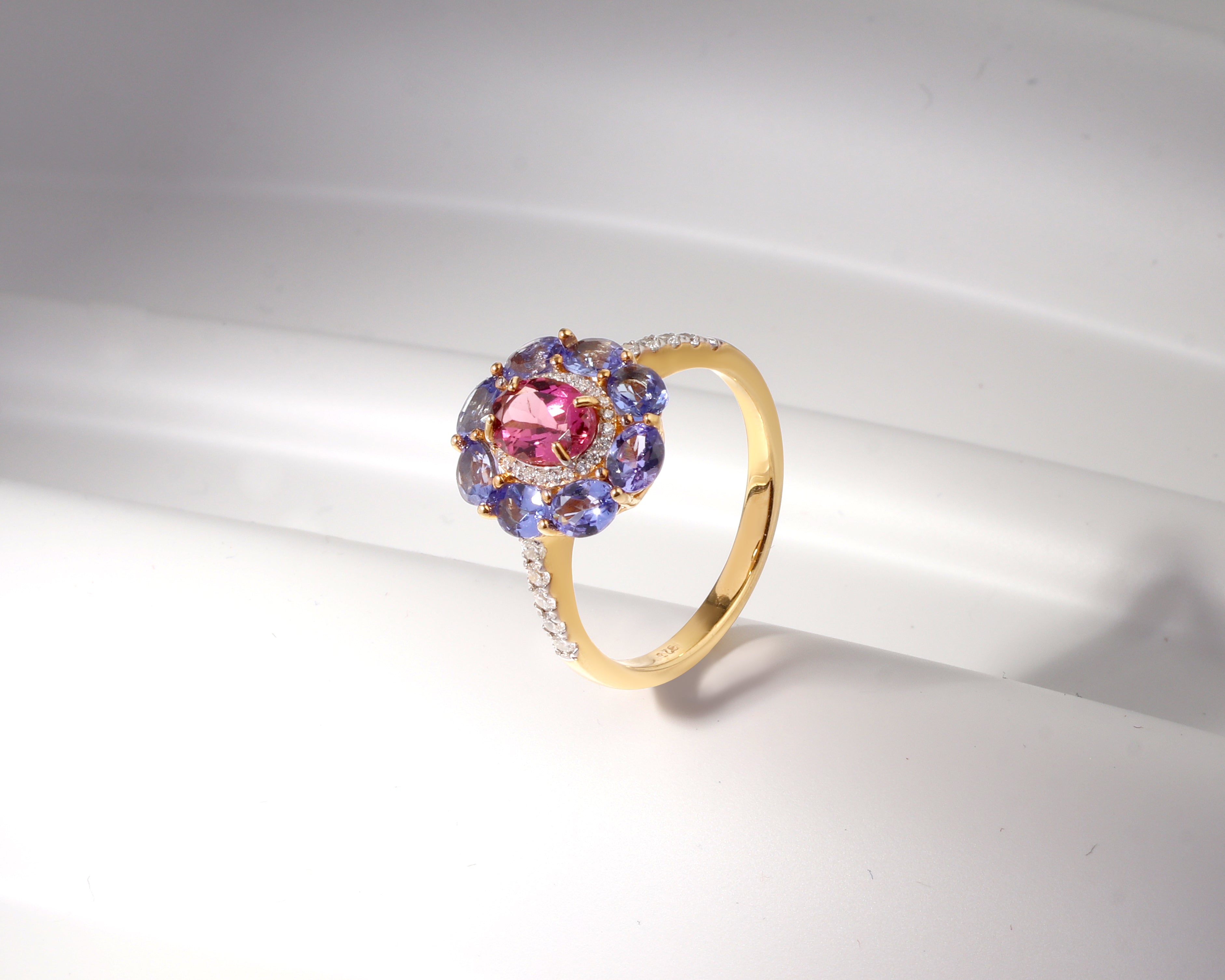 A yellow gold ring featuring a central oval pink gemstone surrounded by a halo of round blue gemstones and small clear accent stones on the band, displayed on a white curved surface.