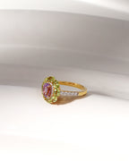 A yellow gold ring with a central oval pink gemstone surrounded by a halo of light green gemstones, and a slim band accented with small clear stones, displayed on a white surface.