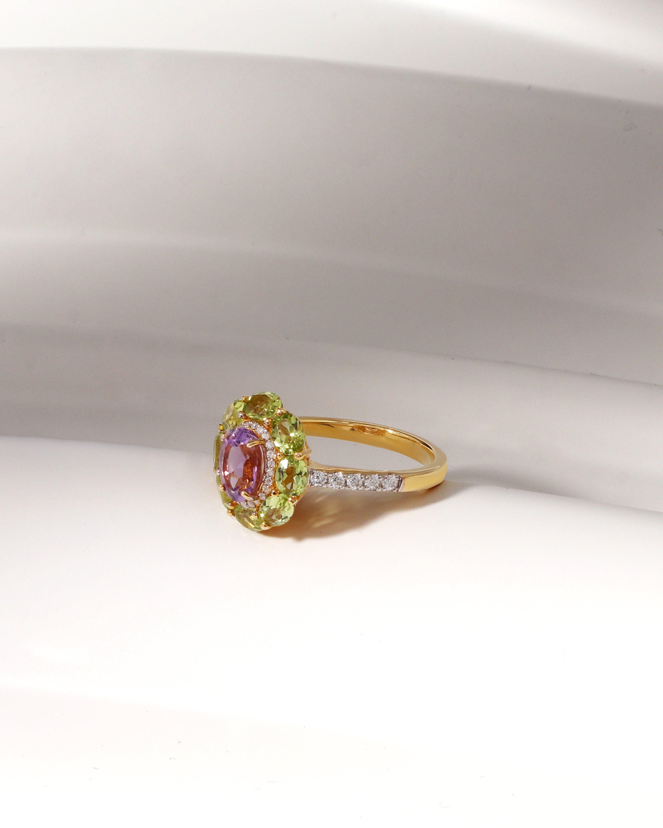 A yellow gold ring with a central oval pink gemstone surrounded by a halo of light green gemstones, and a slim band accented with small clear stones, displayed on a white surface.