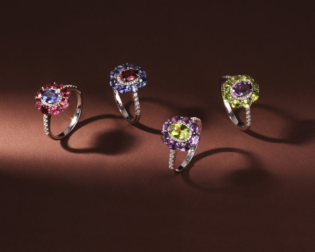 Four silver rings with halo gemstone designs are displayed on a dark brown background. 
