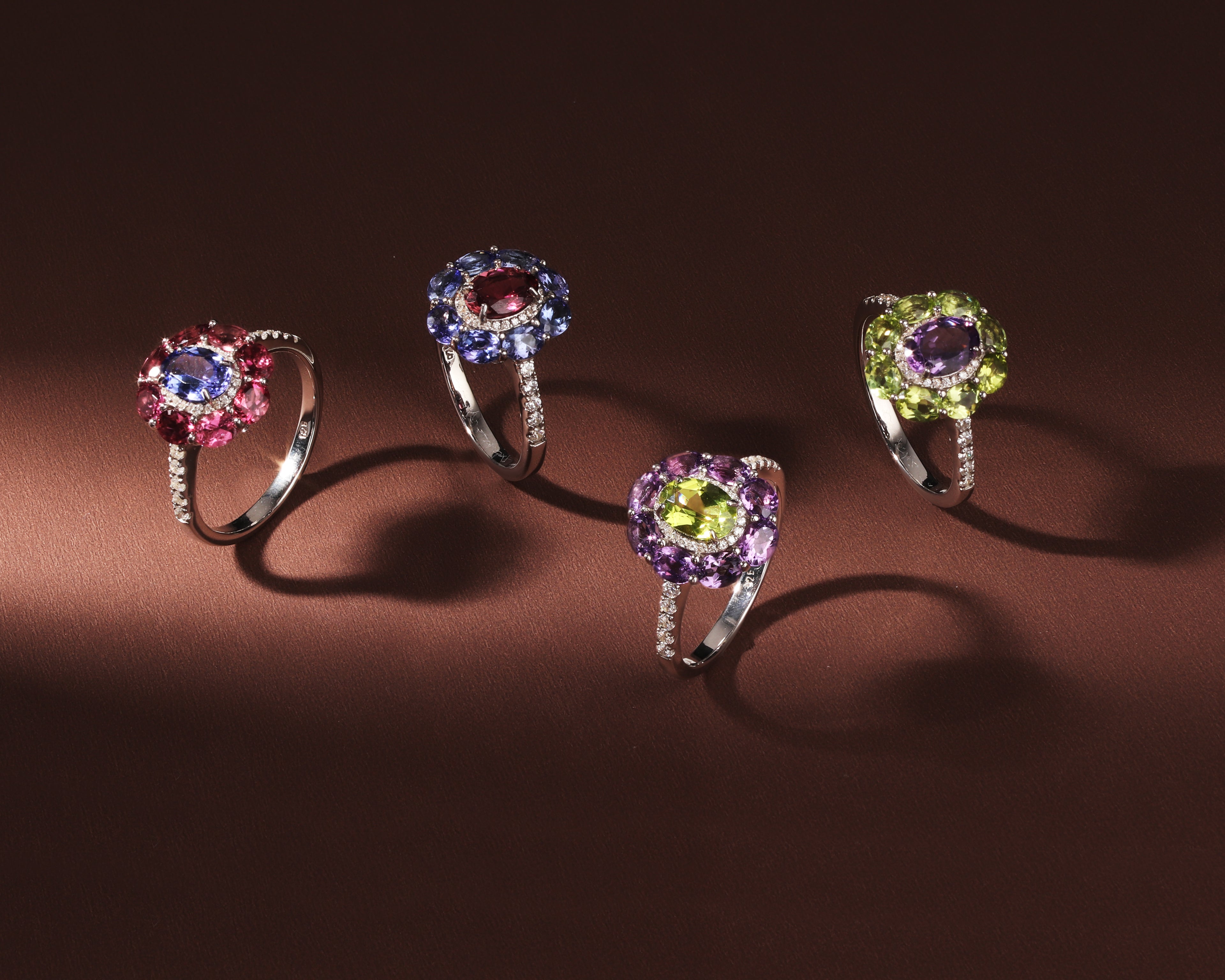 Four silver rings with halo gemstone designs are displayed on a dark brown background. 