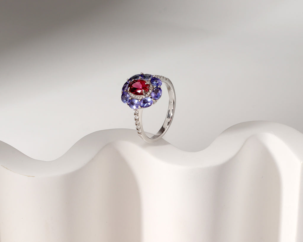 A silver ring featuring a central oval red gemstone surrounded by a halo of round blue gemstones, displayed on a white wavy stand against a light gray background.