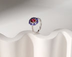 A silver ring featuring a central oval red gemstone surrounded by a halo of round blue gemstones, displayed on a white wavy stand against a light gray background.