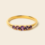 A yellow gold ring featuring five round-cut purple gemstones set across the front, photographed on a clean, solid light beige background.