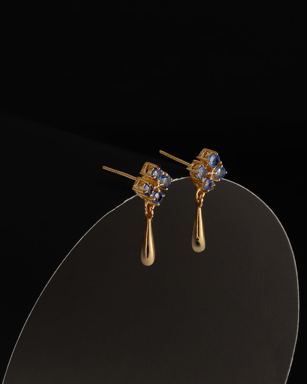 A pair of gold drop earrings with small blue gemstone clusters at the top and teardrop-shaped gold drops below, displayed on a curved clear stand against a black background.