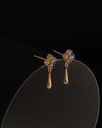 A pair of gold drop earrings with small blue gemstone clusters at the top and teardrop-shaped gold drops below, displayed on a curved clear stand against a black background.
