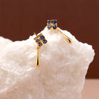 A pair of gold drop earrings with small clusters of dark blue gemstones at the top and teardrop-shaped gold drops below, displayed on a white textured stone against a warm brown background.