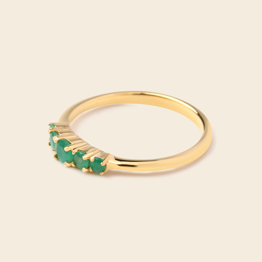 A gold ring featuring a row of small green gemstones in a prong setting, shown on a plain beige background.
