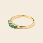 A gold ring featuring a row of small green gemstones in a prong setting, shown on a plain beige background.