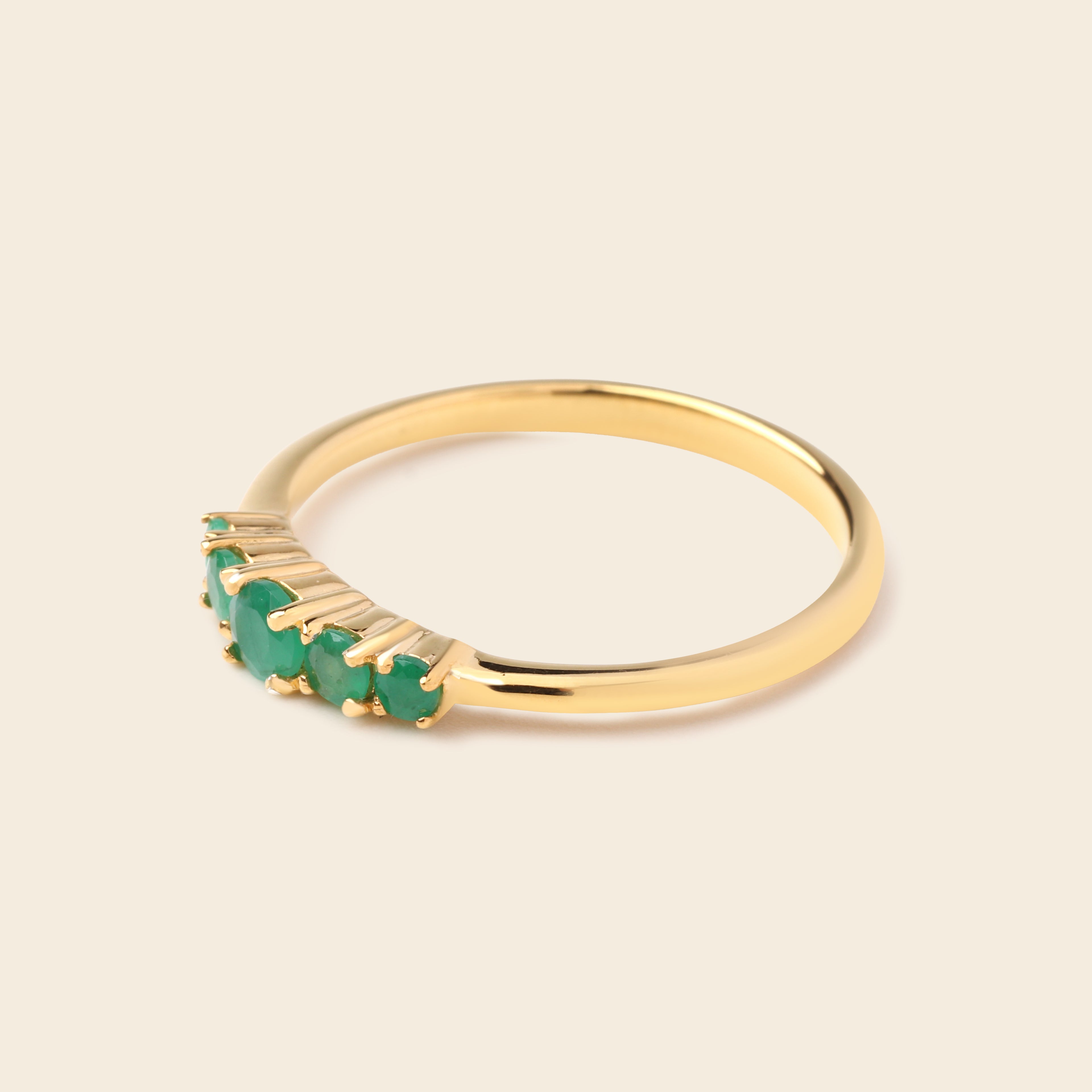 A gold ring featuring a row of small green gemstones in a prong setting, shown on a plain beige background.