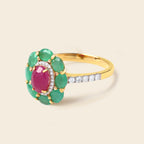 A yellow gold ring featuring a central oval pinkish-red gemstone surrounded by a halo of green oval gemstones, with a pavé band of small clear stones, set against a light beige background.