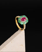A yellow gold ring with a flower-shaped cluster setting featuring green emerald-cut stones and a central oval pinkish-red ruby center stone, accented with small clear pavé stones, displayed on a light-colored stone block against a black background.