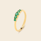 A gold ring featuring five round-cut green gemstones set in a row along the upper band, shown on a light beige background.