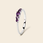 A silver ring featuring a curved row of four oval purple gemstones, set against a solid light beige background.