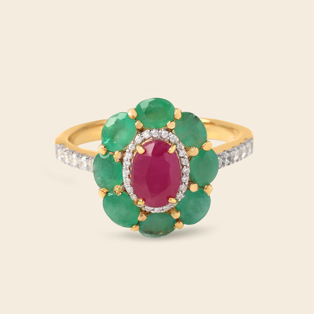 A yellow gold ring featuring a flower-shaped cluster with green emerald-cut stones, a central oval pinkish-red ruby-cut stone, and a halo of small clear stones, set against a light beige background.